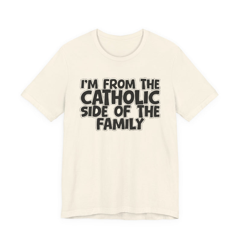 Natural t-shirt with white outlined bubble lettering reading 'I'M FROM THE CATHOLIC SIDE OF THE FAMILY', front flat-lay view.