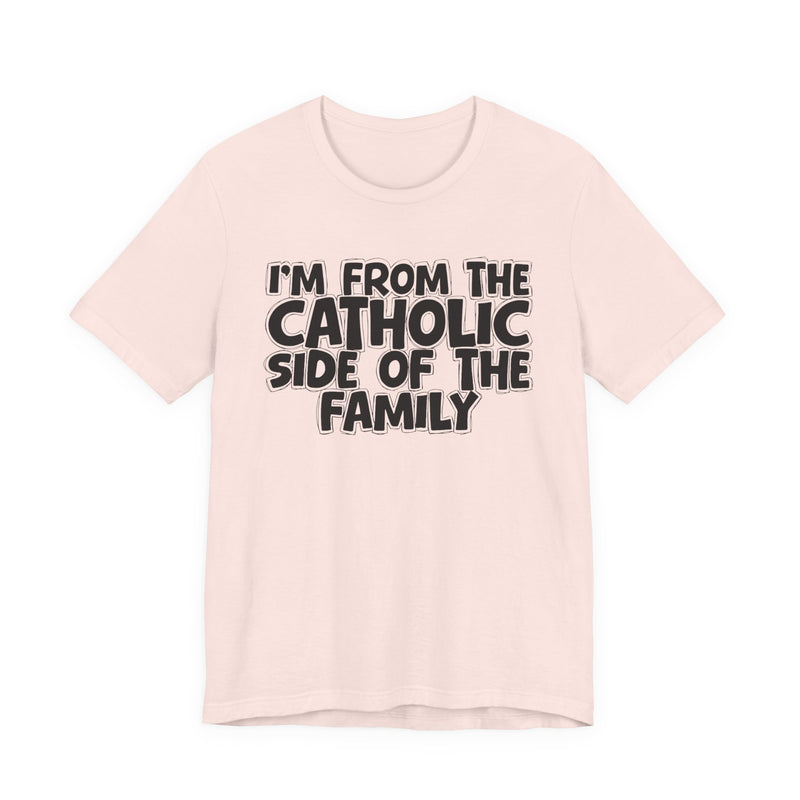 Soft Pink t-shirt with white outlined bubble lettering reading 'I'M FROM THE CATHOLIC SIDE OF THE FAMILY', front flat-lay view.