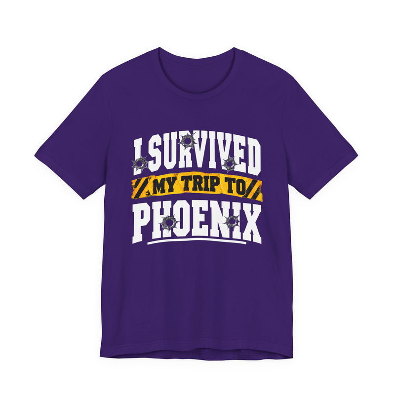 Purple t-shirt with white block lettering reading I SURVIVED MY TRIP TO PHOENIX, with bullet-hole accents and a yellow caution-tape banner, front flat-lay view.