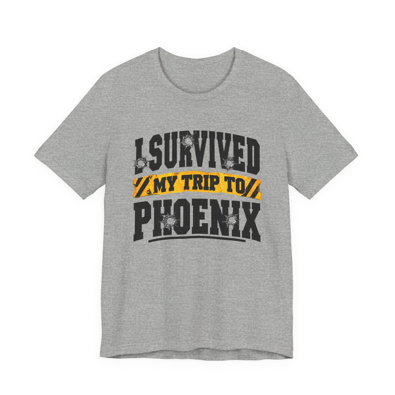 Sport grey t-shirt with black block lettering reading I SURVIVED MY TRIP TO PHOENIX, with bullet-hole accents and a yellow caution-tape banner, front flat-lay view.