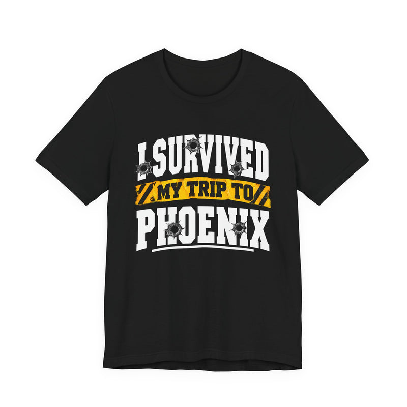Black t-shirt with white block lettering reading I SURVIVED MY TRIP TO PHOENIX, with bullet-hole accents and a yellow caution-tape banner, front flat-lay view.