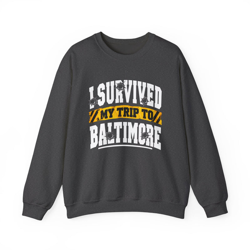 Charcoal sweatshirt with white block lettering reading I SURVIVED MY TRIP TO BALTIMORE, with bullet-hole accents and a yellow caution-tape banner, front flat-lay view.