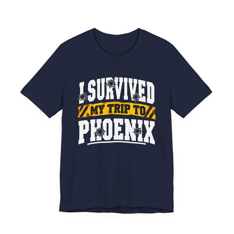 Black t-shirt with white block lettering reading I SURVIVED MY TRIP TO PHOENIX, with bullet-hole accents and a yellow caution-tape banner, front flat-lay view.