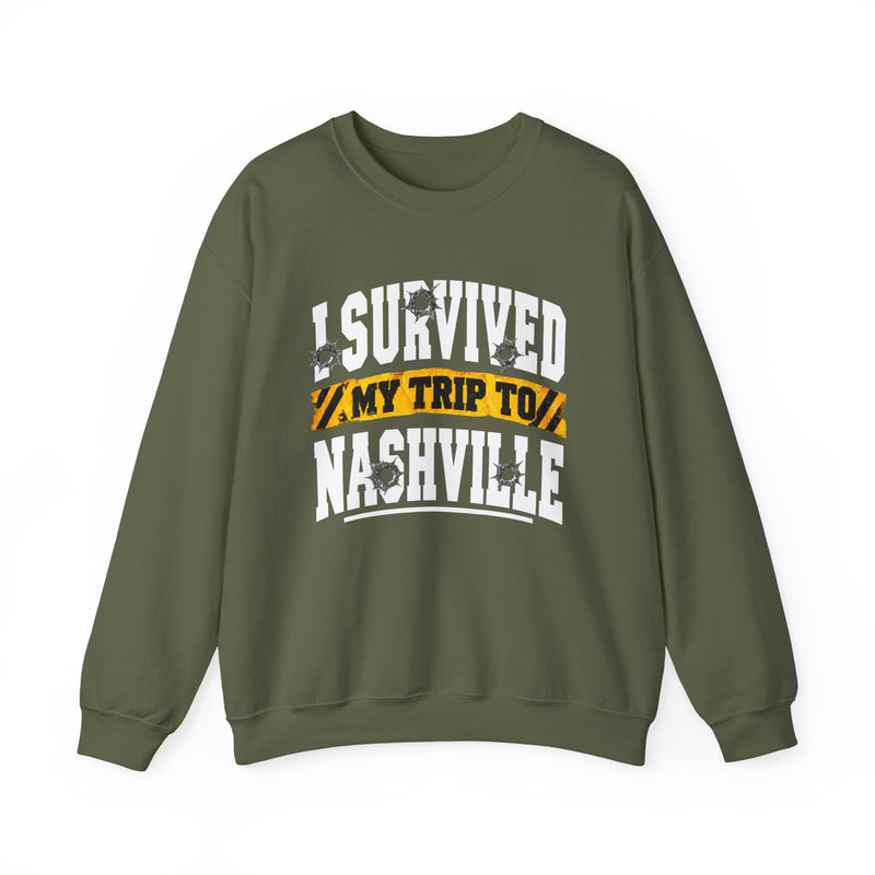 Military green sweatshirt with white block lettering reading I SURVIVED MY TRIP TO NASHVILLE, with bullet-hole accents and a yellow caution-tape banner, front flat-lay view.