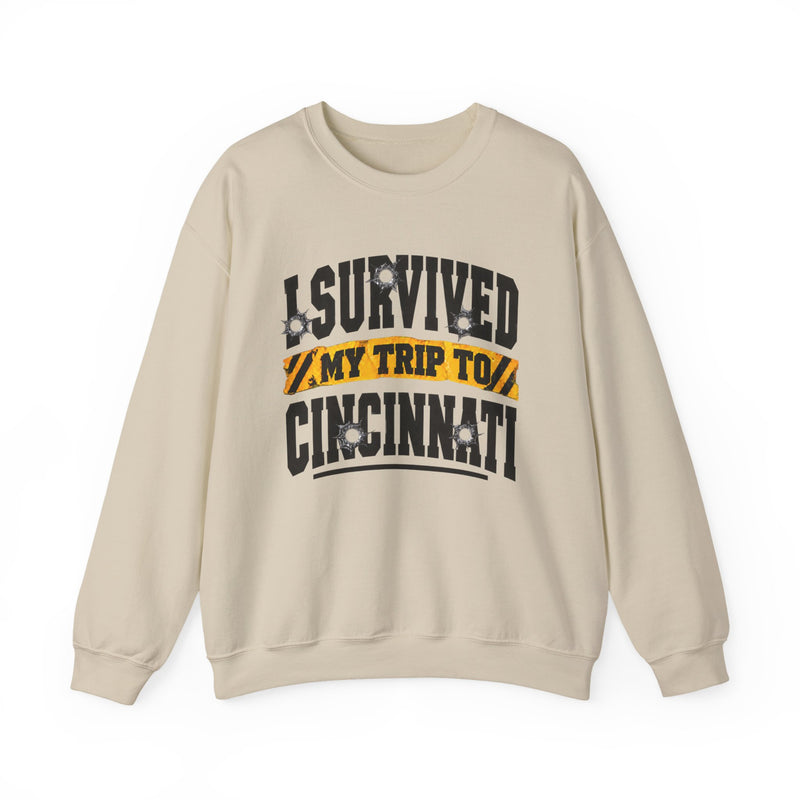 Sand sweatshirt with black block lettering reading I SURVIVED MY TRIP TO CINCINNATI, with bullet-hole accents and a yellow caution-tape banner, front flat-lay view.