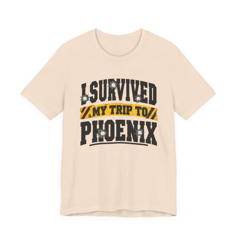 Light pink t-shirt with black block lettering reading I SURVIVED MY TRIP TO PHOENIX, with bullet-hole accents and a yellow caution-tape banner, front flat-lay view.