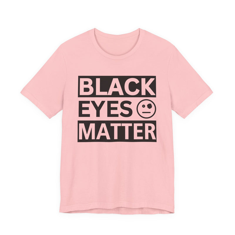 Natural t-shirt with ‘BLACK EYES MATTER’ in boxed block lettering with a smiley-face eye icon graphic in dark-contrast print, front flat-lay mockup view