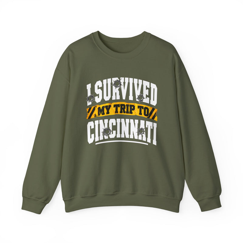 Military green sweatshirt with white block lettering reading I SURVIVED MY TRIP TO CINCINNATI, with bullet-hole accents and a yellow caution-tape banner, front flat-lay view.