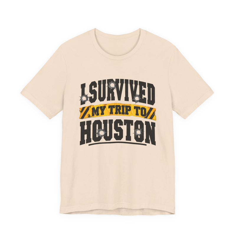 Light pink t-shirt with black block lettering reading I SURVIVED MY TRIP TO HOUSTON, with bullet-hole accents and a yellow caution-tape banner, front flat-lay view.