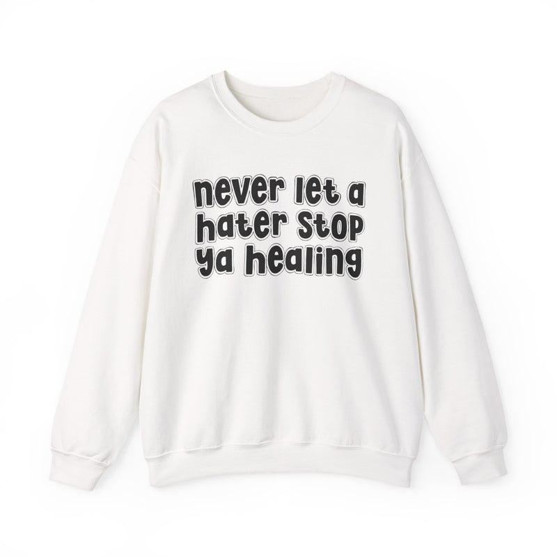 White sweatshirt with dark bold healing-journey lettering reading NEVER LET A HATER STOP YA HEALING, front flat-lay view.