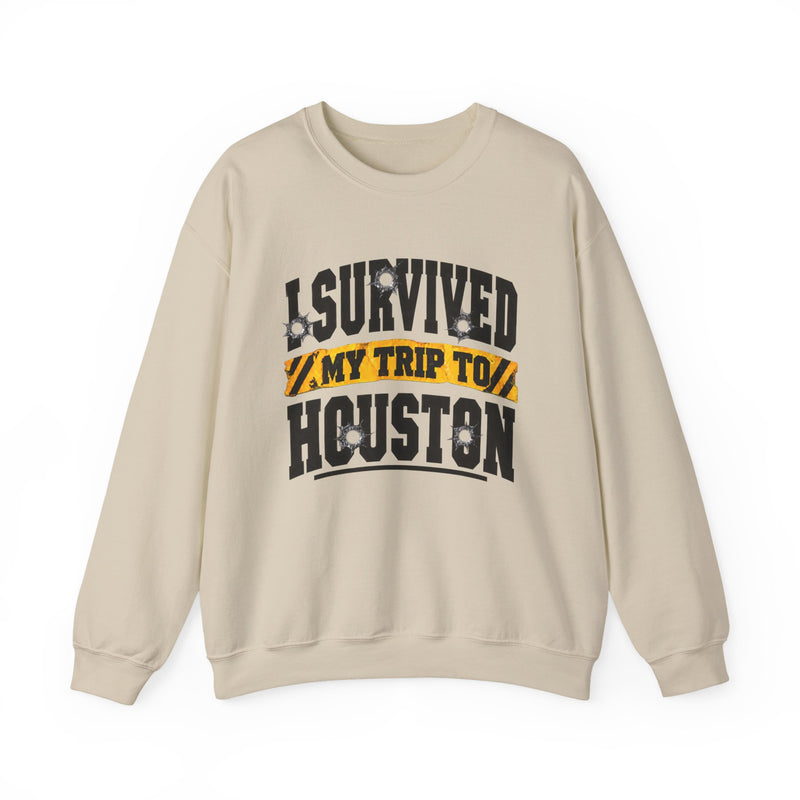 Sand sweatshirt with black block lettering reading I SURVIVED MY TRIP TO HOUSTON, with bullet-hole accents and a yellow caution-tape banner, front flat-lay view.