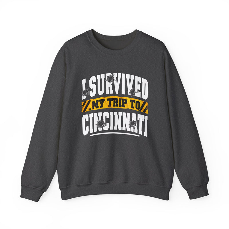 Charcoal sweatshirt with white block lettering reading I SURVIVED MY TRIP TO CINCINNATI, with bullet-hole accents and a yellow caution-tape banner, front flat-lay view.