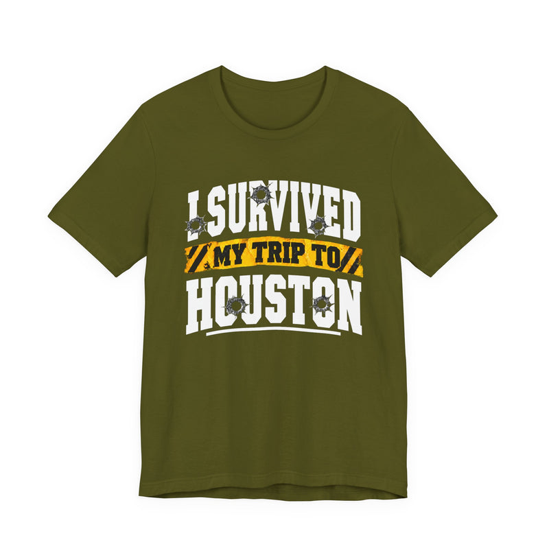 Olive t-shirt with white block lettering reading I SURVIVED MY TRIP TO HOUSTON, with bullet-hole accents and a yellow caution-tape banner, front flat-lay view.