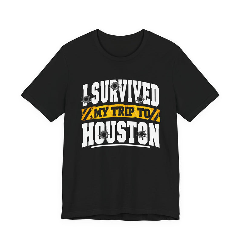 Black t-shirt with white block lettering reading I SURVIVED MY TRIP TO HOUSTON, with bullet-hole accents and a yellow caution-tape banner, front flat-lay view.