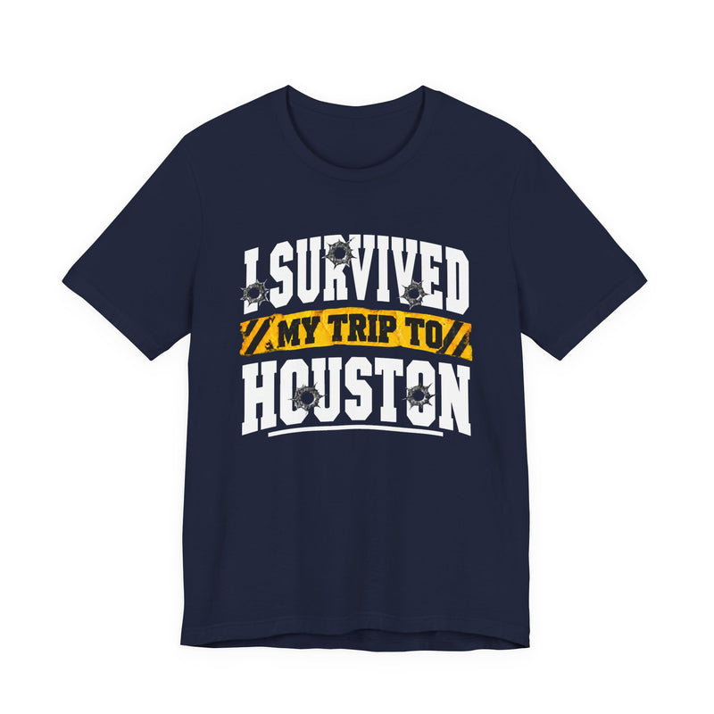 Black t-shirt with white block lettering reading I SURVIVED MY TRIP TO HOUSTON, with bullet-hole accents and a yellow caution-tape banner, front flat-lay view.