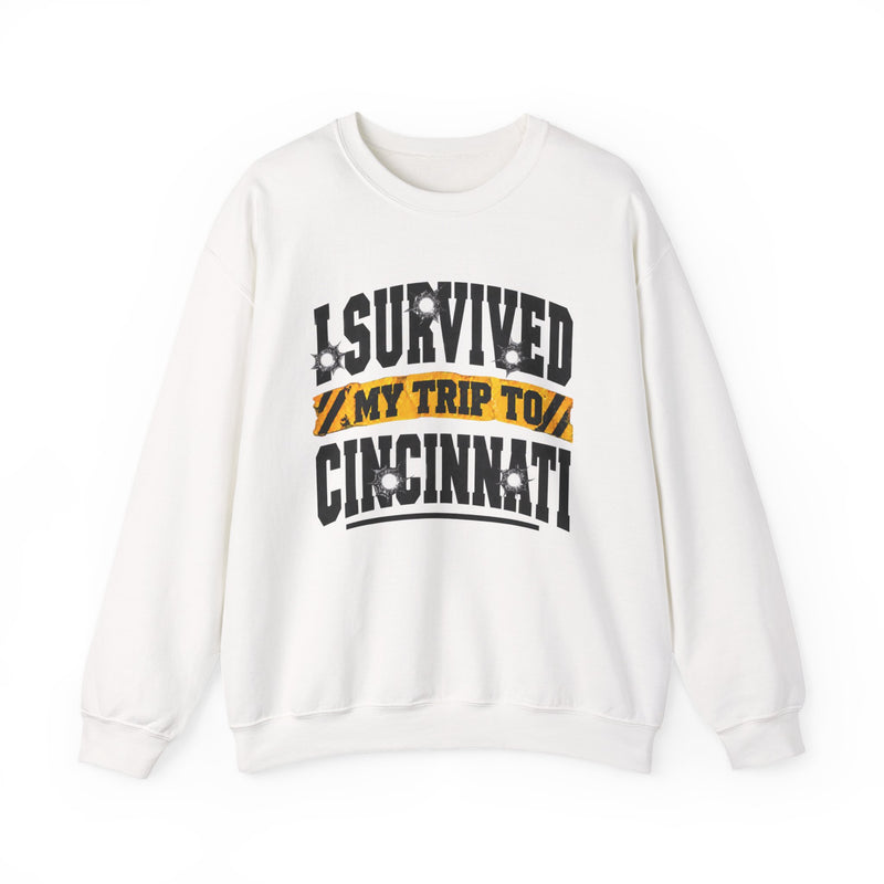 White sweatshirt with black block lettering reading I SURVIVED MY TRIP TO CINCINNATI, with bullet-hole accents and a yellow caution-tape banner, front flat-lay view.