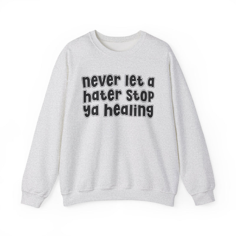 Natural sweatshirt with dark bold healing-journey lettering reading NEVER LET A HATER STOP YA HEALING, front flat-lay view.