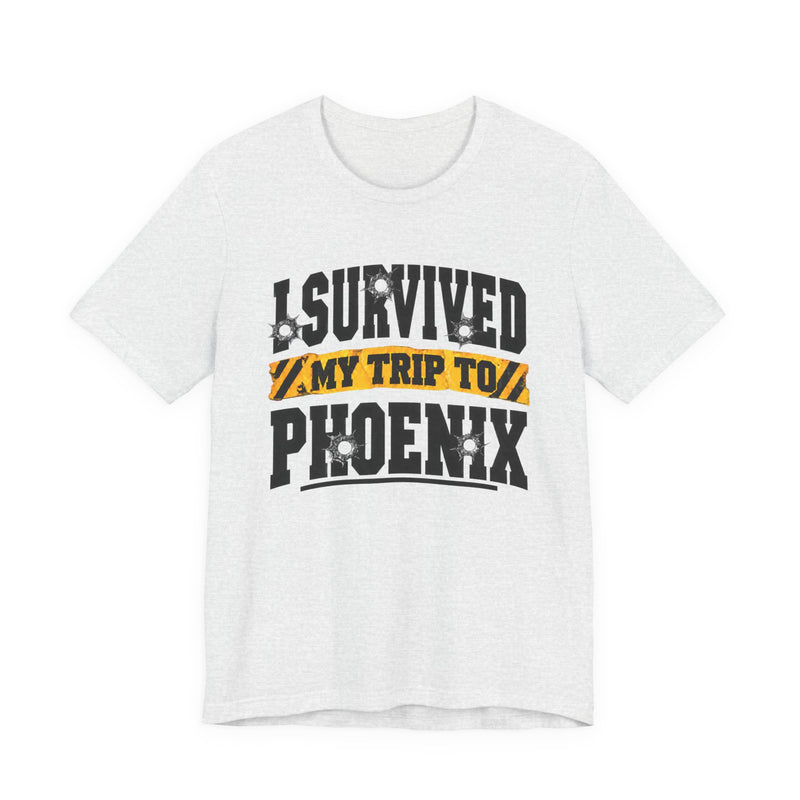 Black t-shirt with white block lettering reading I SURVIVED MY TRIP TO PHOENIX, with bullet-hole accents and a yellow caution-tape banner, front flat-lay view.
