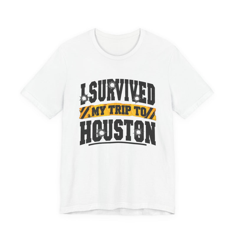 Black t-shirt with white block lettering reading I SURVIVED MY TRIP TO HOUSTON, with bullet-hole accents and a yellow caution-tape banner, front flat-lay view.