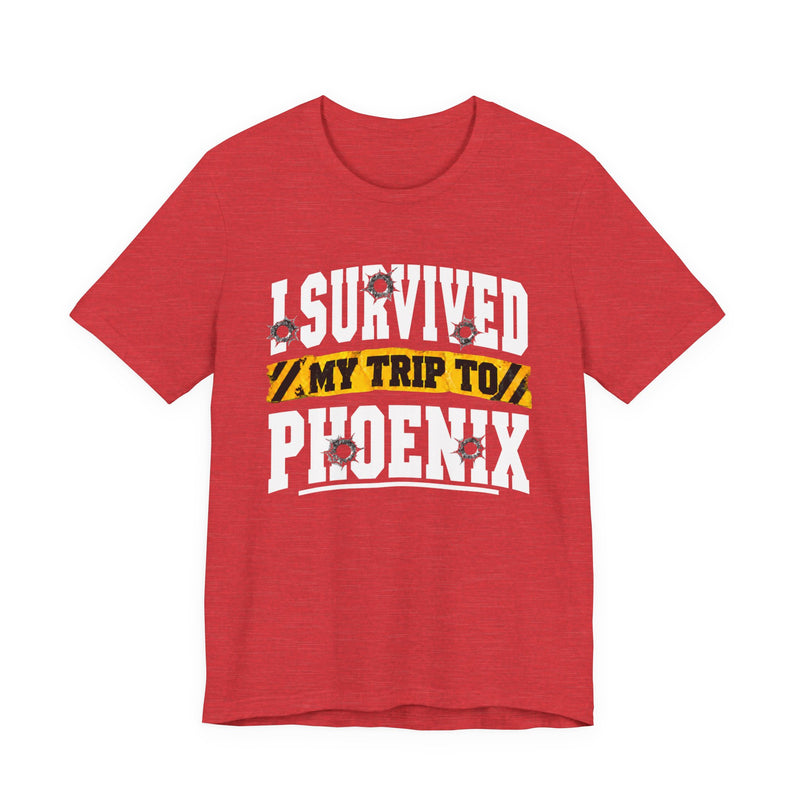 Red t-shirt with white block lettering reading I SURVIVED MY TRIP TO PHOENIX, with bullet-hole accents and a yellow caution-tape banner, front flat-lay view.