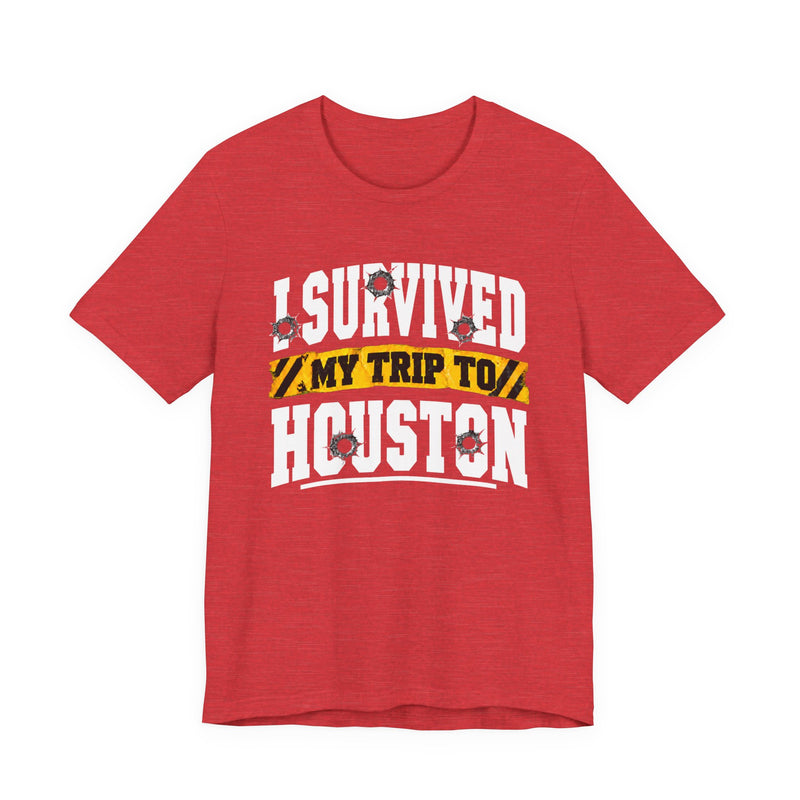 Red t-shirt with white block lettering reading I SURVIVED MY TRIP TO HOUSTON, with bullet-hole accents and a yellow caution-tape banner, front flat-lay view.