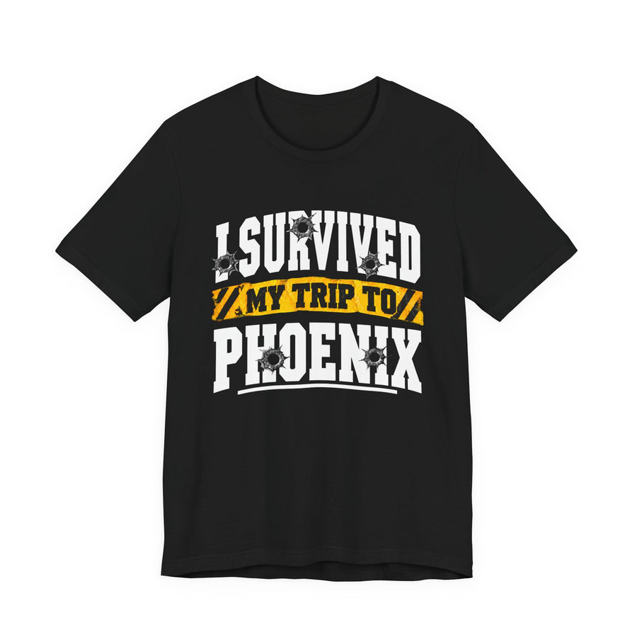 Black t-shirt with white block lettering reading I SURVIVED MY TRIP TO PHOENIX, with bullet-hole accents and a yellow caution-tape banner, front flat-lay view.