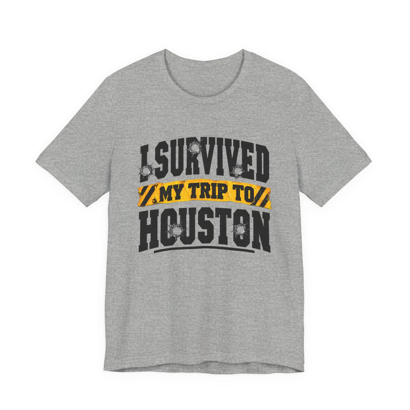 Sport grey t-shirt with black block lettering reading I SURVIVED MY TRIP TO HOUSTON, with bullet-hole accents and a yellow caution-tape banner, front flat-lay view.