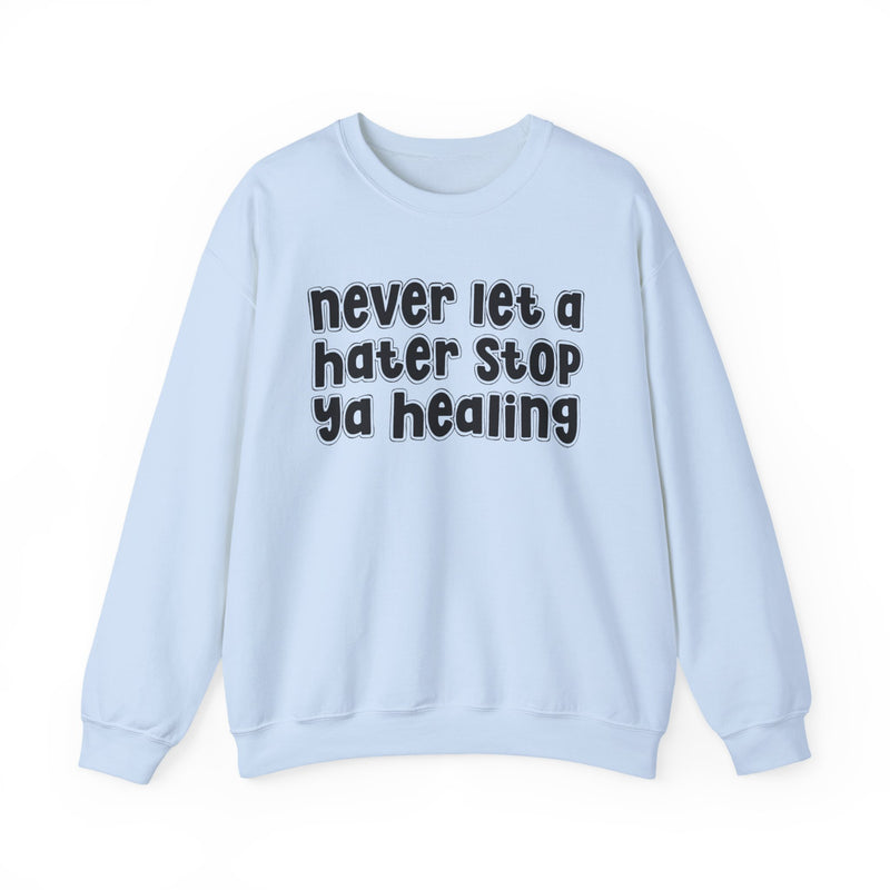 White sweatshirt with dark bold healing-journey lettering reading NEVER LET A HATER STOP YA HEALING, front flat-lay view.