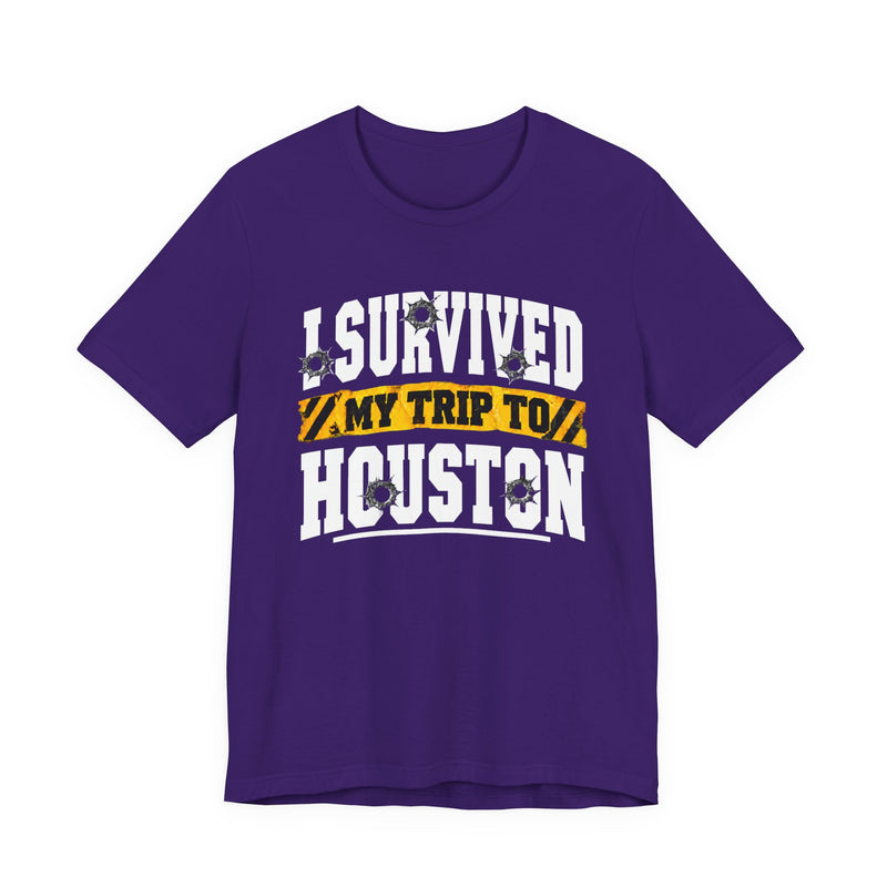 Purple t-shirt with white block lettering reading I SURVIVED MY TRIP TO HOUSTON, with bullet-hole accents and a yellow caution-tape banner, front flat-lay view.