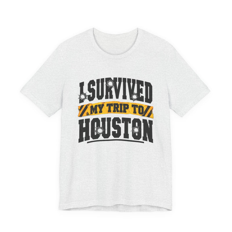 Black t-shirt with white block lettering reading I SURVIVED MY TRIP TO HOUSTON, with bullet-hole accents and a yellow caution-tape banner, front flat-lay view.