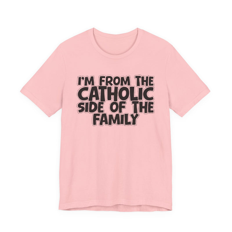 Pink t-shirt with white outlined bubble lettering reading 'I'M FROM THE CATHOLIC SIDE OF THE FAMILY', front flat-lay view.