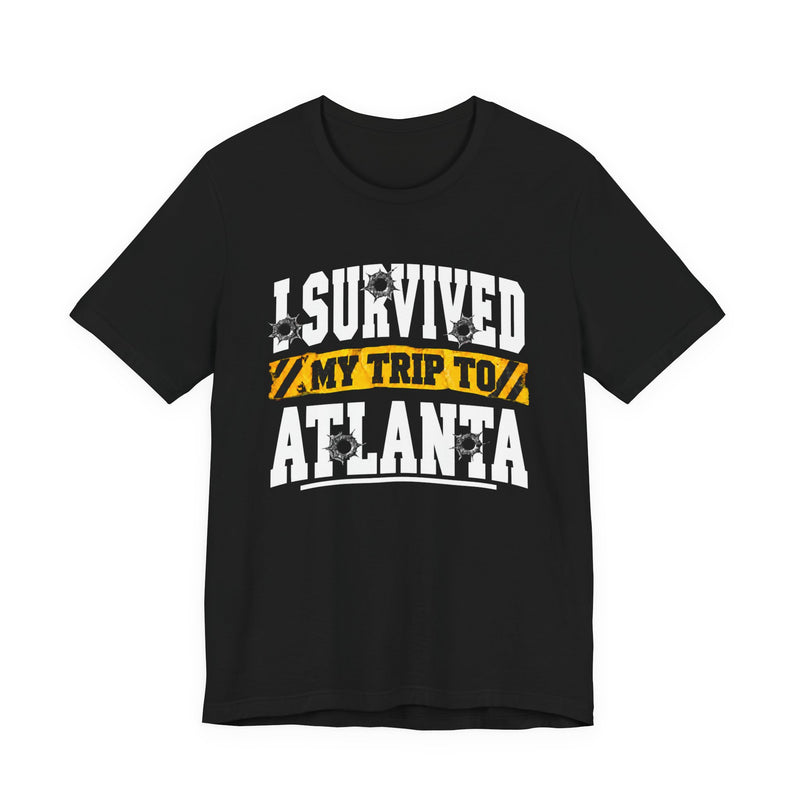 Black t-shirt with white collegiate lettering reading I SURVIVED MY TRIP TO ATLANTA with yellow caution-tape banner and bullet-hole graphics, front flat-lay view.