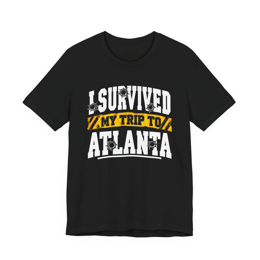Black t-shirt with white collegiate lettering reading I SURVIVED MY TRIP TO ATLANTA with yellow caution-tape banner and bullet-hole graphics, front flat-lay view.