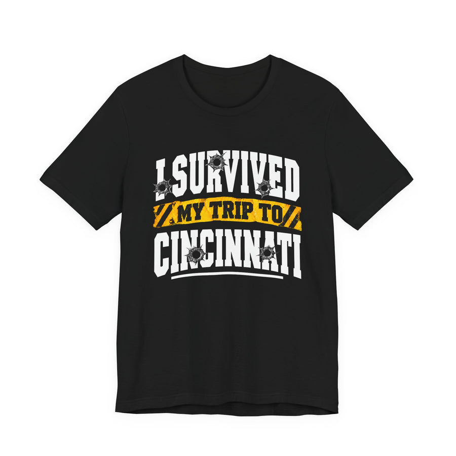 Black t-shirt with white block lettering reading I SURVIVED MY TRIP TO CINCINNATI, with bullet-hole accents and a yellow caution-tape banner, front flat-lay view.