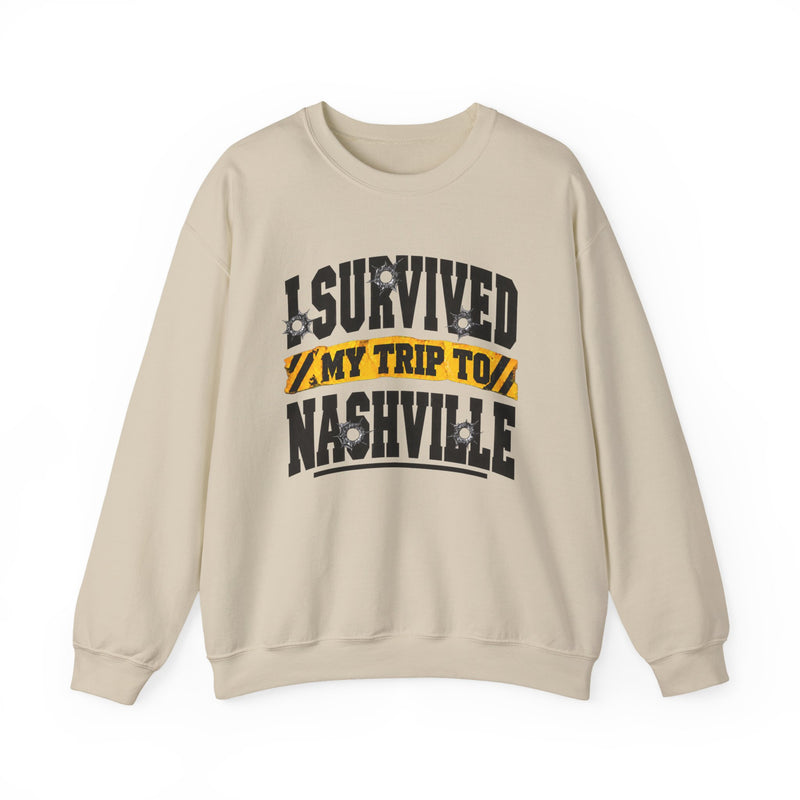 Sand sweatshirt with black block lettering reading I SURVIVED MY TRIP TO NASHVILLE, with bullet-hole accents and a yellow caution-tape banner, front flat-lay view.