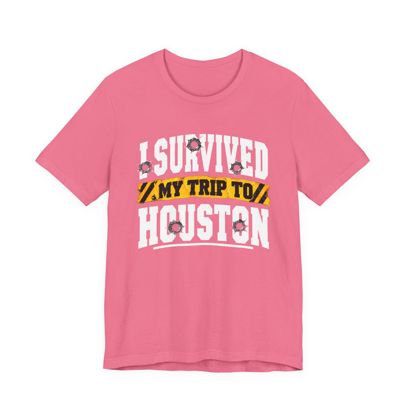 Hot pink t-shirt with white block lettering reading I SURVIVED MY TRIP TO HOUSTON, with bullet-hole accents and a yellow caution-tape banner, front flat-lay view.