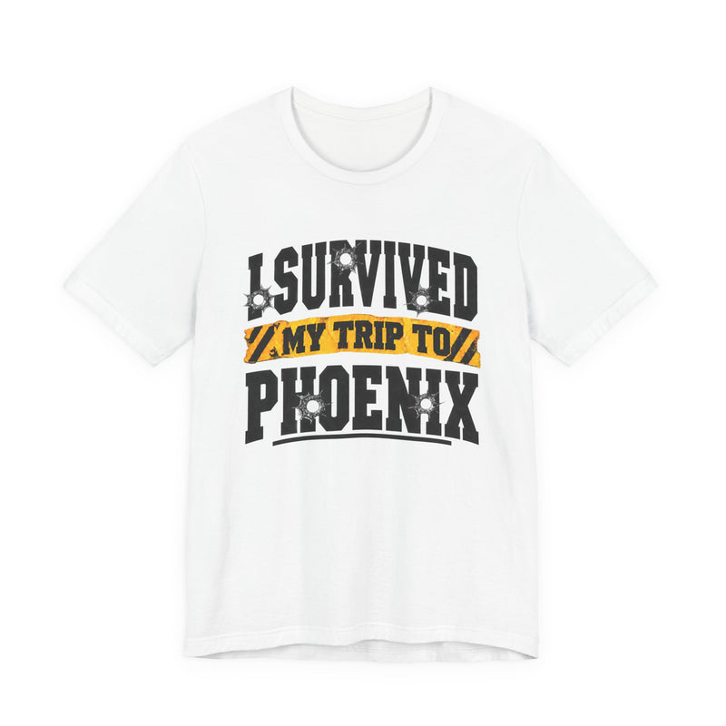 Black t-shirt with white block lettering reading I SURVIVED MY TRIP TO PHOENIX, with bullet-hole accents and a yellow caution-tape banner, front flat-lay view.