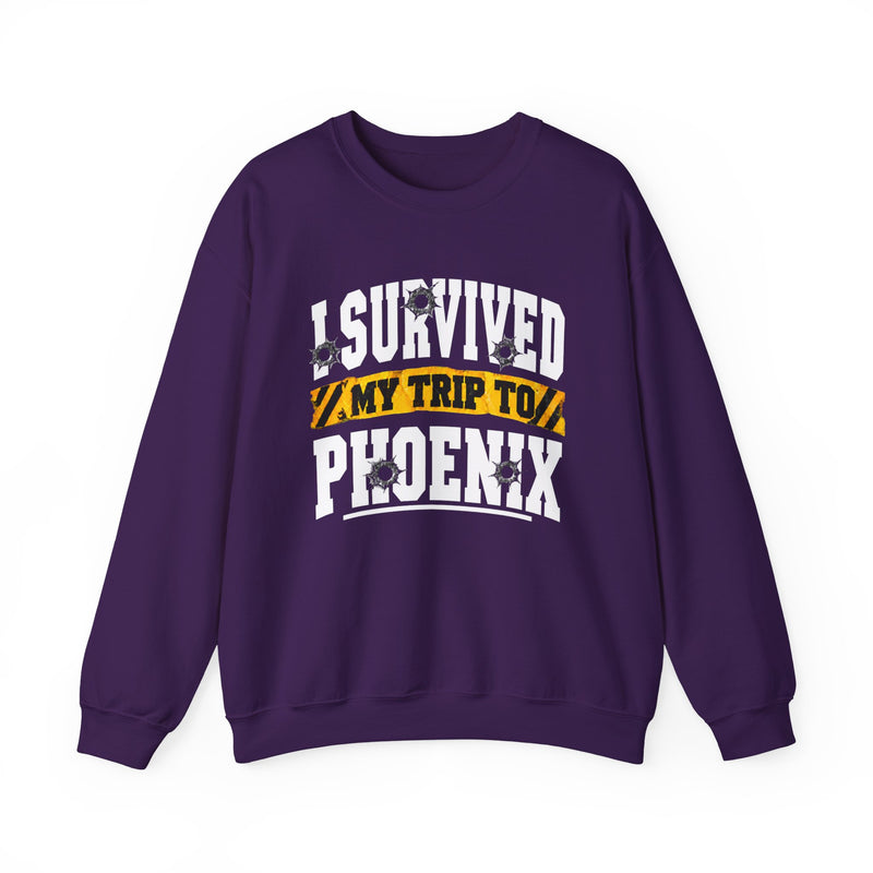 Purple sweatshirt with white block lettering reading I SURVIVED MY TRIP TO PHOENIX, with bullet-hole accents and a yellow caution-tape banner, front flat-lay view.