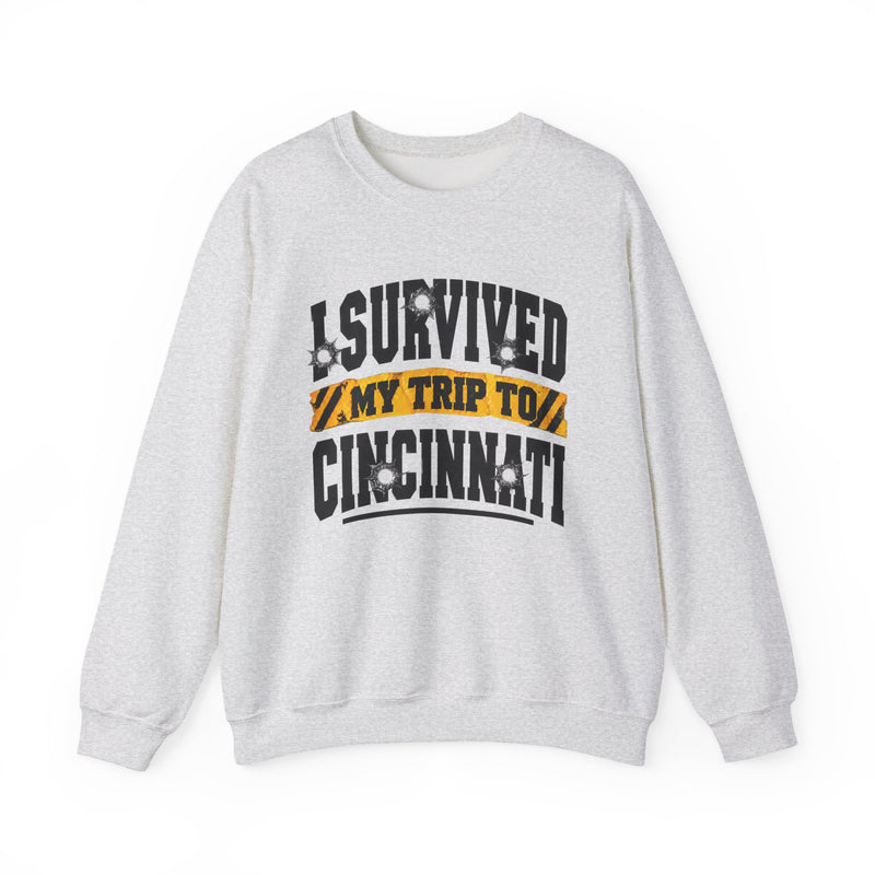 White sweatshirt with black block lettering reading I SURVIVED MY TRIP TO CINCINNATI, with bullet-hole accents and a yellow caution-tape banner, front flat-lay view.