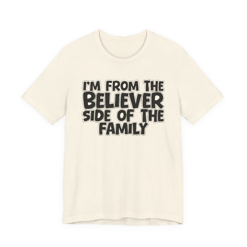 Natural t-shirt with white outlined bubble lettering reading 'I'M FROM THE BELIEVER SIDE OF THE FAMILY', front flat-lay view.