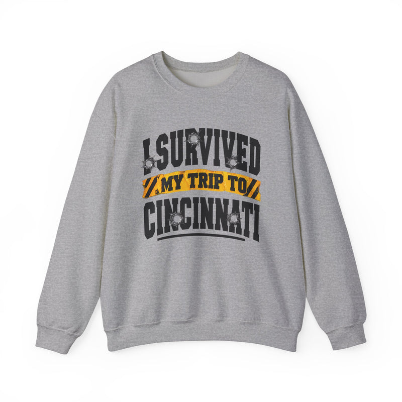 Sport grey sweatshirt with black block lettering reading I SURVIVED MY TRIP TO CINCINNATI, with bullet-hole accents and a yellow caution-tape banner, front flat-lay view.