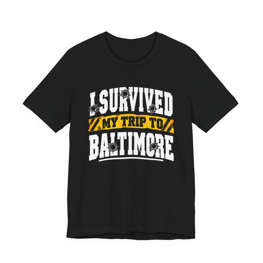 Black t-shirt with white block lettering reading I SURVIVED MY TRIP TO BALTIMORE, with bullet-hole accents and a yellow caution-tape banner, front flat-lay view.