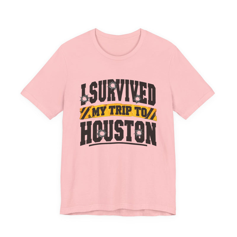 Light pink t-shirt with black block lettering reading I SURVIVED MY TRIP TO HOUSTON, with bullet-hole accents and a yellow caution-tape banner, front flat-lay view.