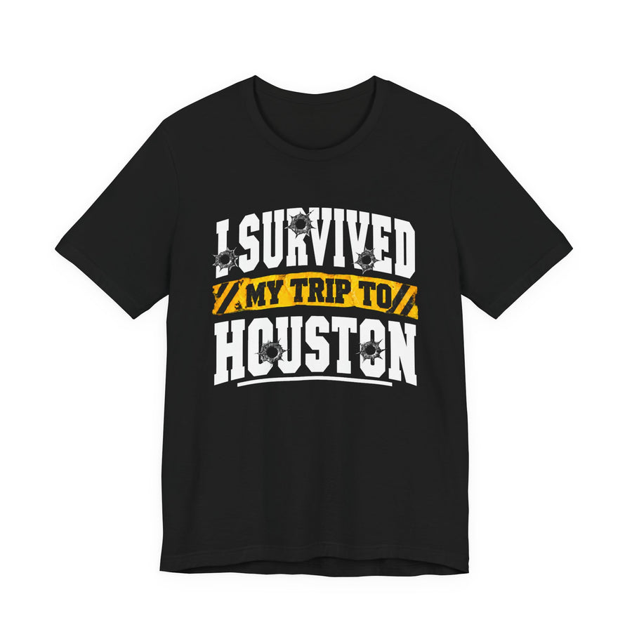 Black t-shirt with white block lettering reading I SURVIVED MY TRIP TO HOUSTON, with bullet-hole accents and a yellow caution-tape banner, front flat-lay view.