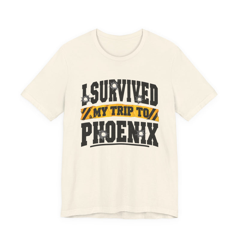 White t-shirt with black block lettering reading I SURVIVED MY TRIP TO PHOENIX, with bullet-hole accents and a yellow caution-tape banner, front flat-lay view.