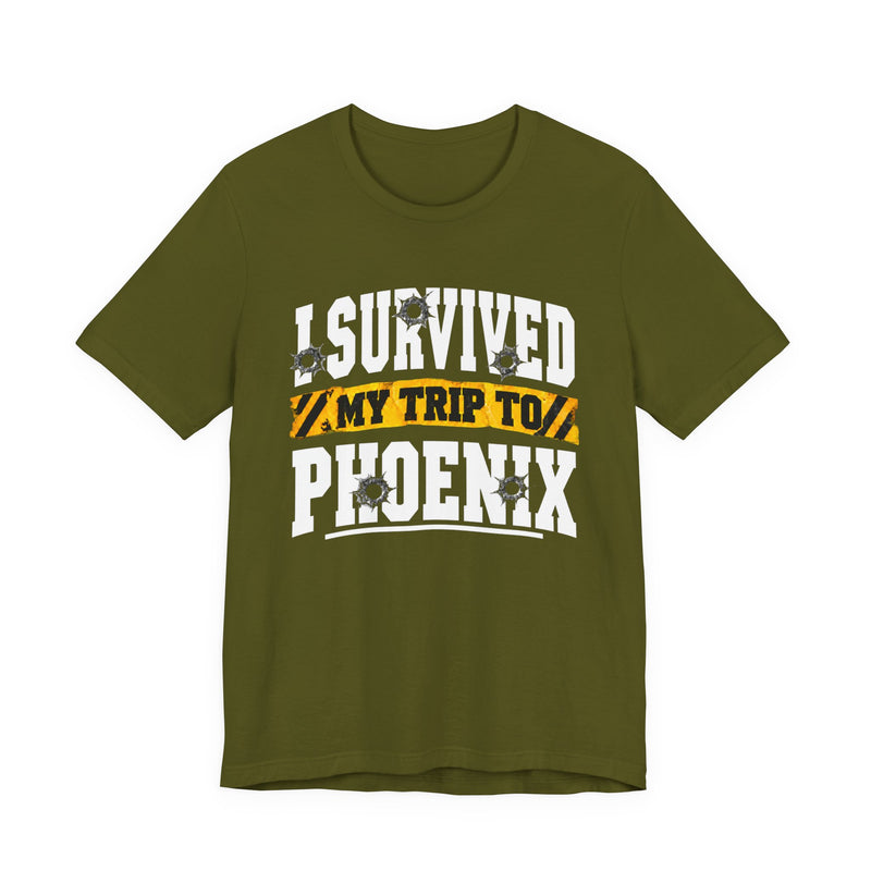 Olive t-shirt with white block lettering reading I SURVIVED MY TRIP TO PHOENIX, with bullet-hole accents and a yellow caution-tape banner, front flat-lay view.