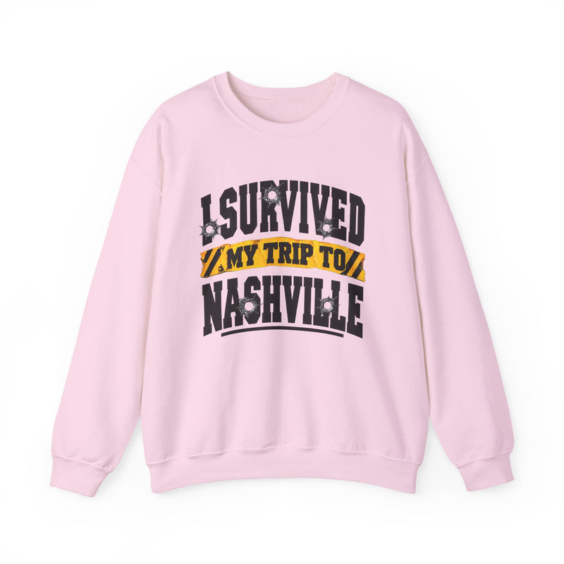 Light pink sweatshirt with black block lettering reading I SURVIVED MY TRIP TO NASHVILLE, with bullet-hole accents and a yellow caution-tape banner, front flat-lay view.