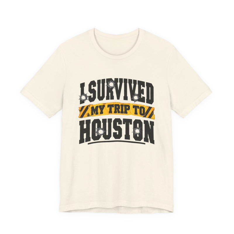 White t-shirt with black block lettering reading I SURVIVED MY TRIP TO HOUSTON, with bullet-hole accents and a yellow caution-tape banner, front flat-lay view.