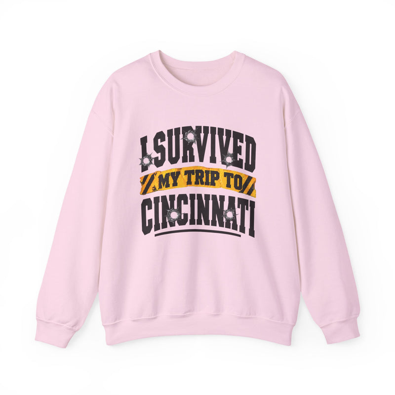 Light pink sweatshirt with black block lettering reading I SURVIVED MY TRIP TO CINCINNATI, with bullet-hole accents and a yellow caution-tape banner, front flat-lay view.