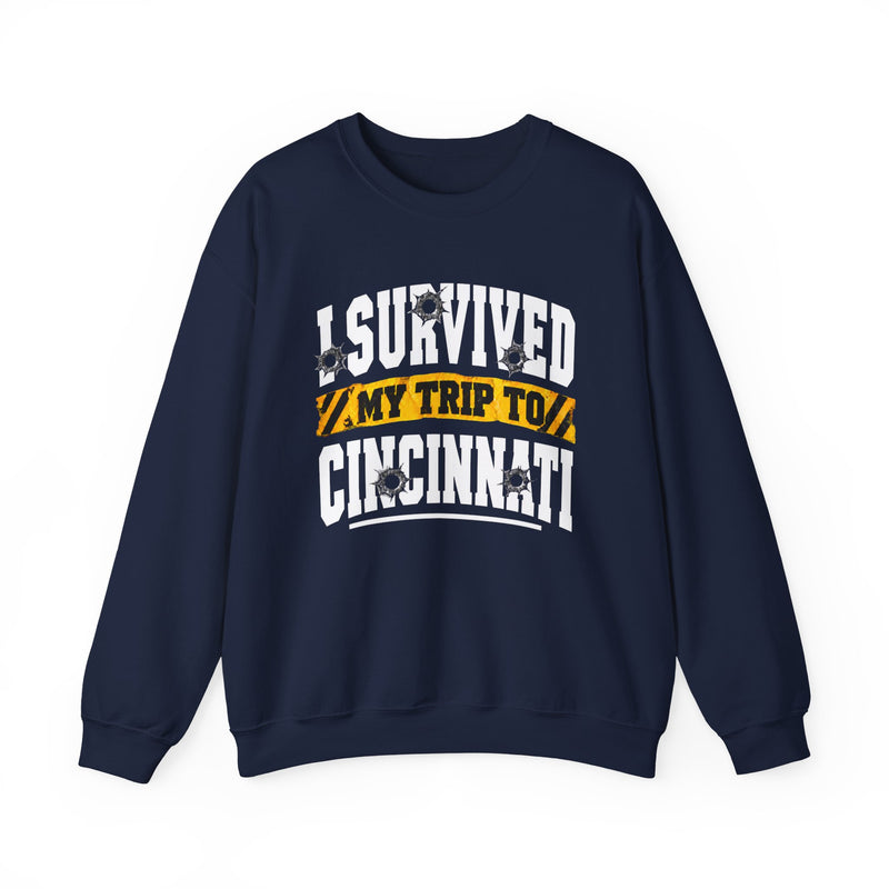 Navy sweatshirt with white block lettering reading I SURVIVED MY TRIP TO CINCINNATI, with bullet-hole accents and a yellow caution-tape banner, front flat-lay view.